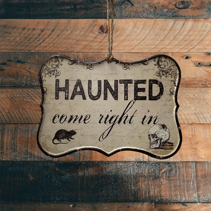 Hanging Sign that says Haunted come right in and has an image of a skull and a rat in the bottom corners. The Sign is hanging from a wooden wall 