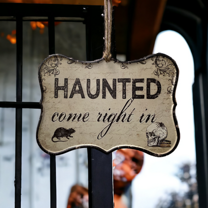 Hanging Sign that says Haunted come right in and has an image of a skull and a rat in the bottom corners. The Sign is hanging from a black  frame and glass door