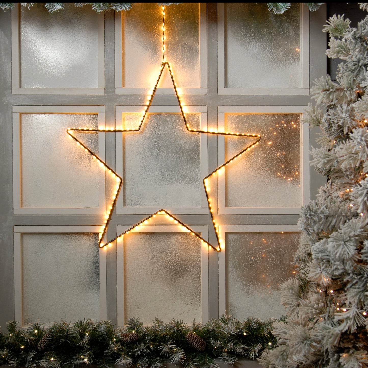 lit star hanging in front of a snowy window with pinecone green garland and a snowy garland surrounding the window 