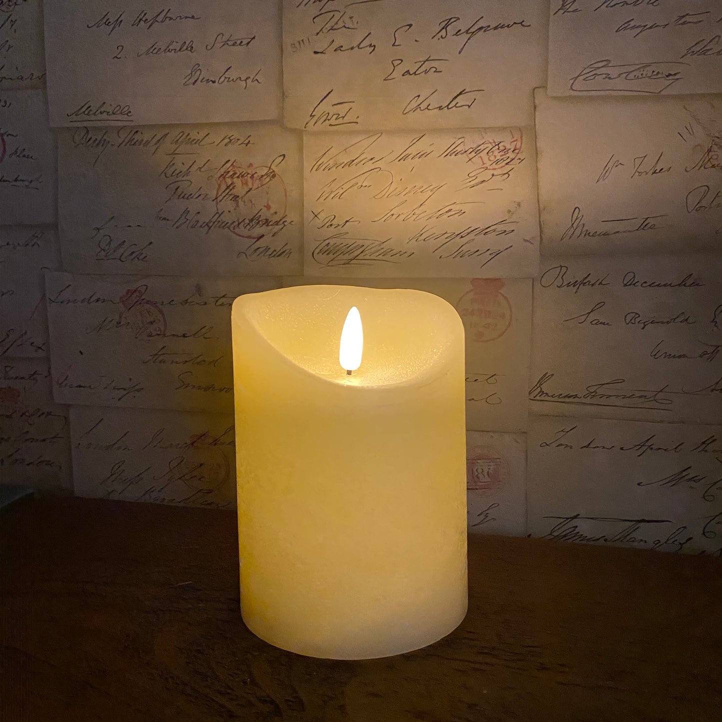 cream candle with the LED on, on top of a wooden mantle with a envelope design wall paper in the background