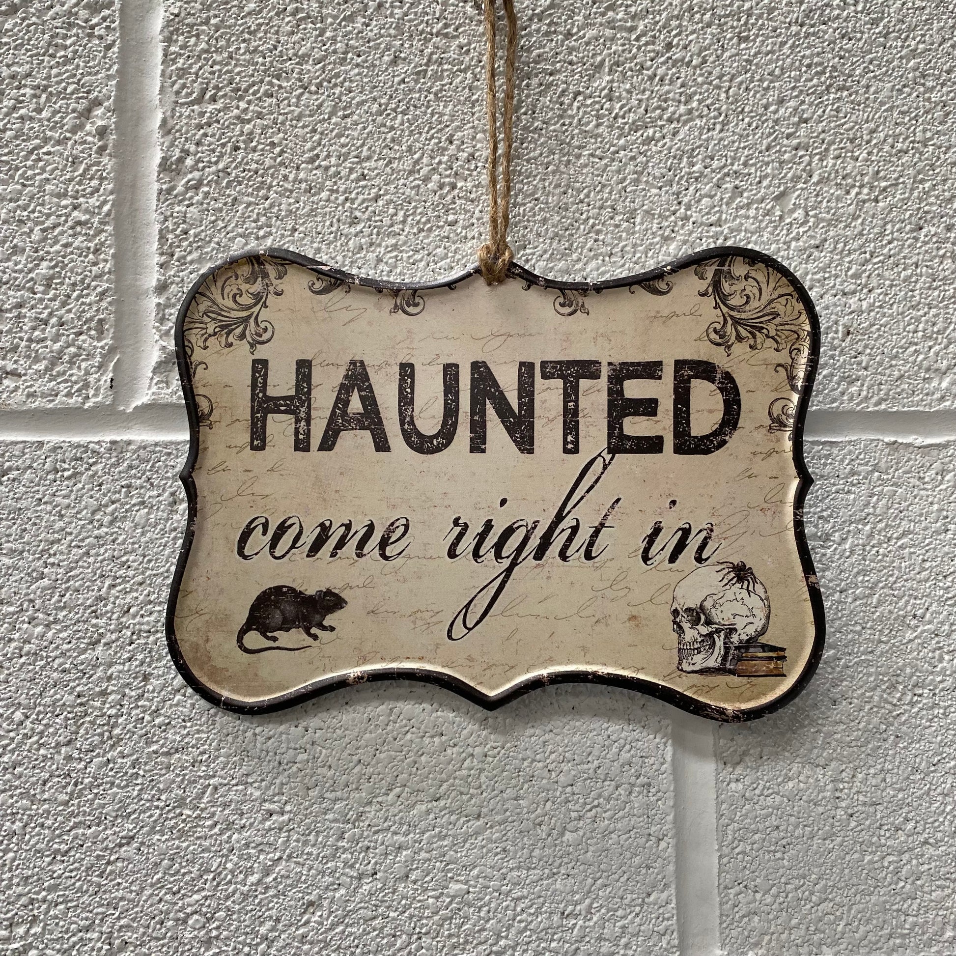 Hanging Sign that says Haunted come right in and has an image of a skull and a rat in the bottom corners. The Sign is hanging from a white brick wall